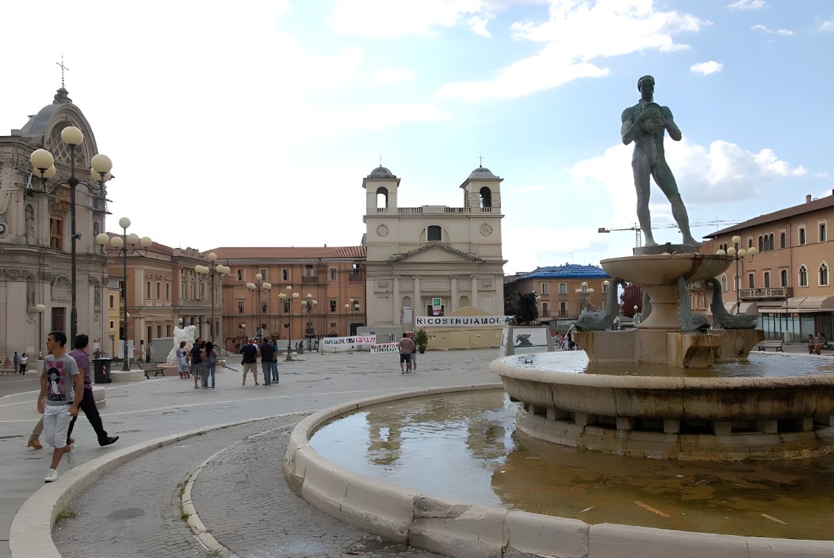 Pedestrians walk through Piazza Duomo in L'Aquila past the Fontana Vecchia and street lighting poles.