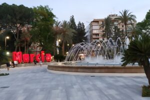 Large red Murcia sign next to a circular public fountain with arched water jets in a city plaza at dusk.