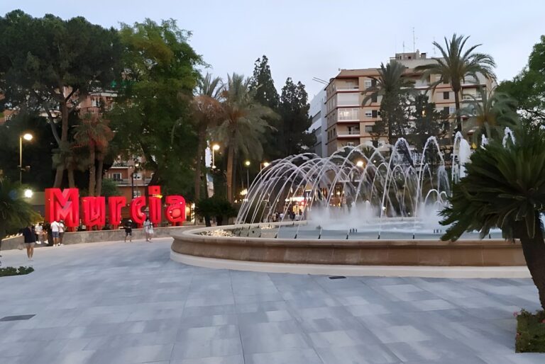 Large red Murcia sign next to a circular public fountain with arched water jets in a city plaza at dusk.