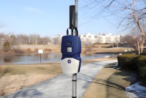 An N5 Sensors wildfire detection device mounted on a metal pole in a park with a pond and walking path in the background.