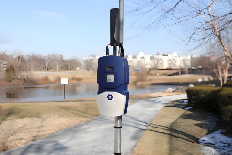 An N5 Sensors wildfire detection device mounted on a metal pole in a park with a pond and walking path in the background.