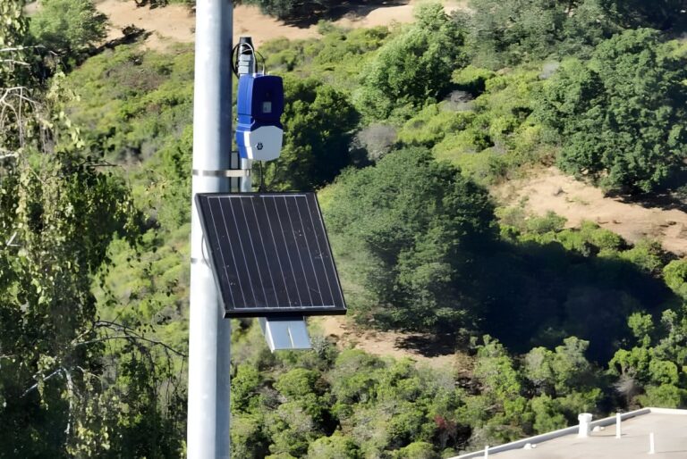 An N5 Sensors wildfire detection device and solar panel mounted on a utility pole overlooking a wooded hillside.