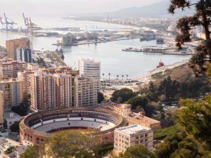 High-angle view of the Málaga bullring and city skyline overlooking the industrial port and Mediterranean coastline.