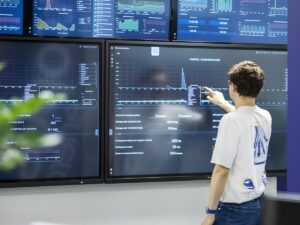 A technician points at a data visualization dashboard on a large wall-mounted monitor in a control room.