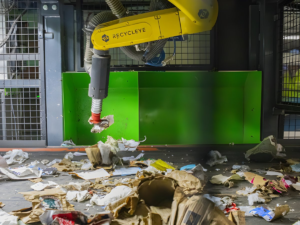 A yellow Recycleye robotic arm sorts paper and cardboard waste on a conveyor belt in a recycling facility.