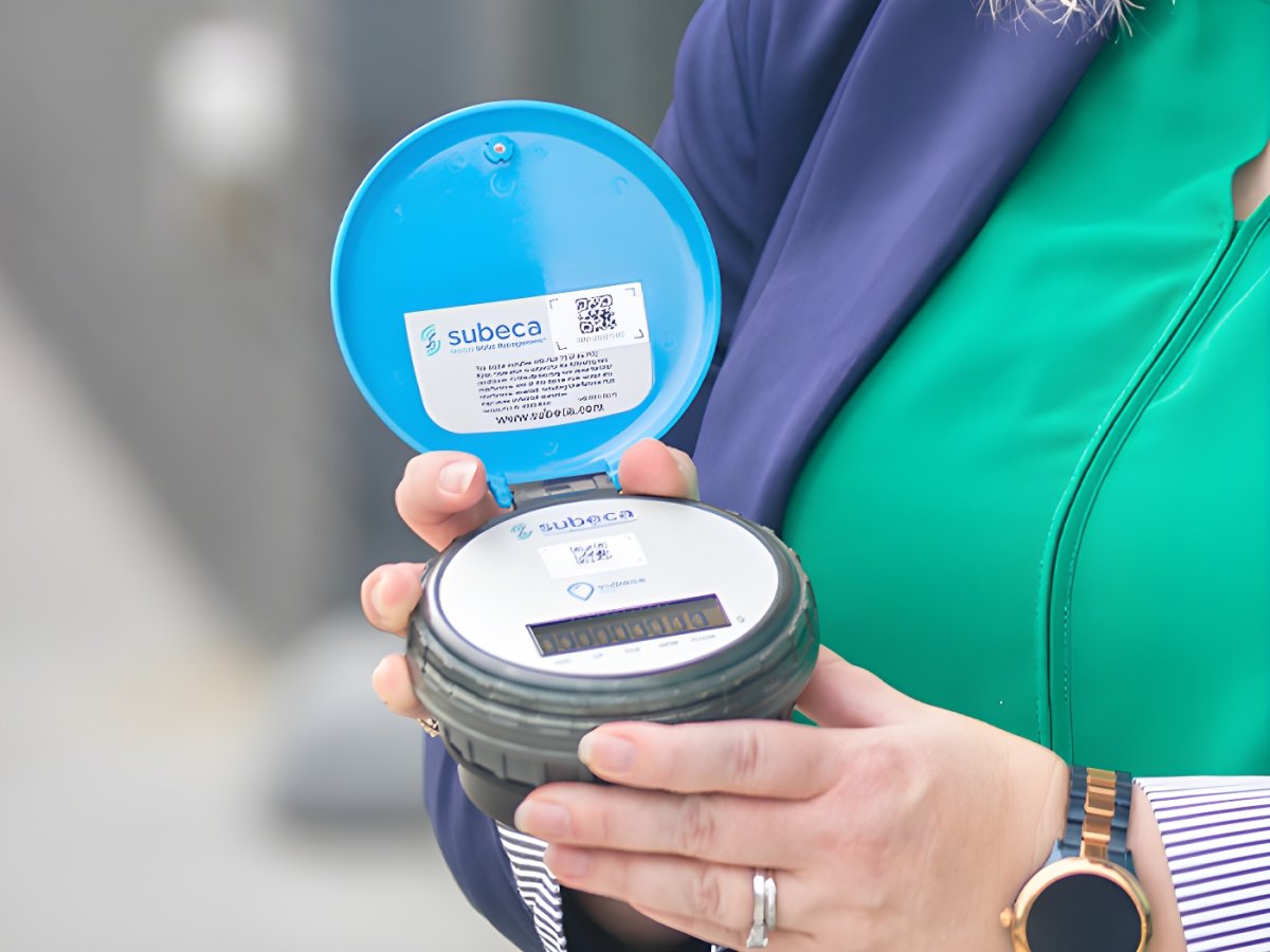 A person holding a Subeca smart water meter with an open blue lid and digital display.
