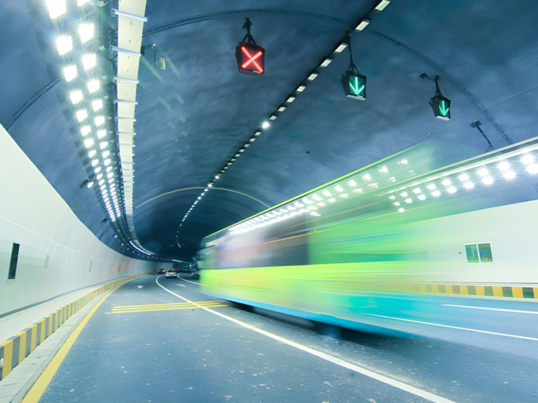 Motion blur of a green bus driving through a brightly lit highway tunnel with overhead digital lane control signals.
