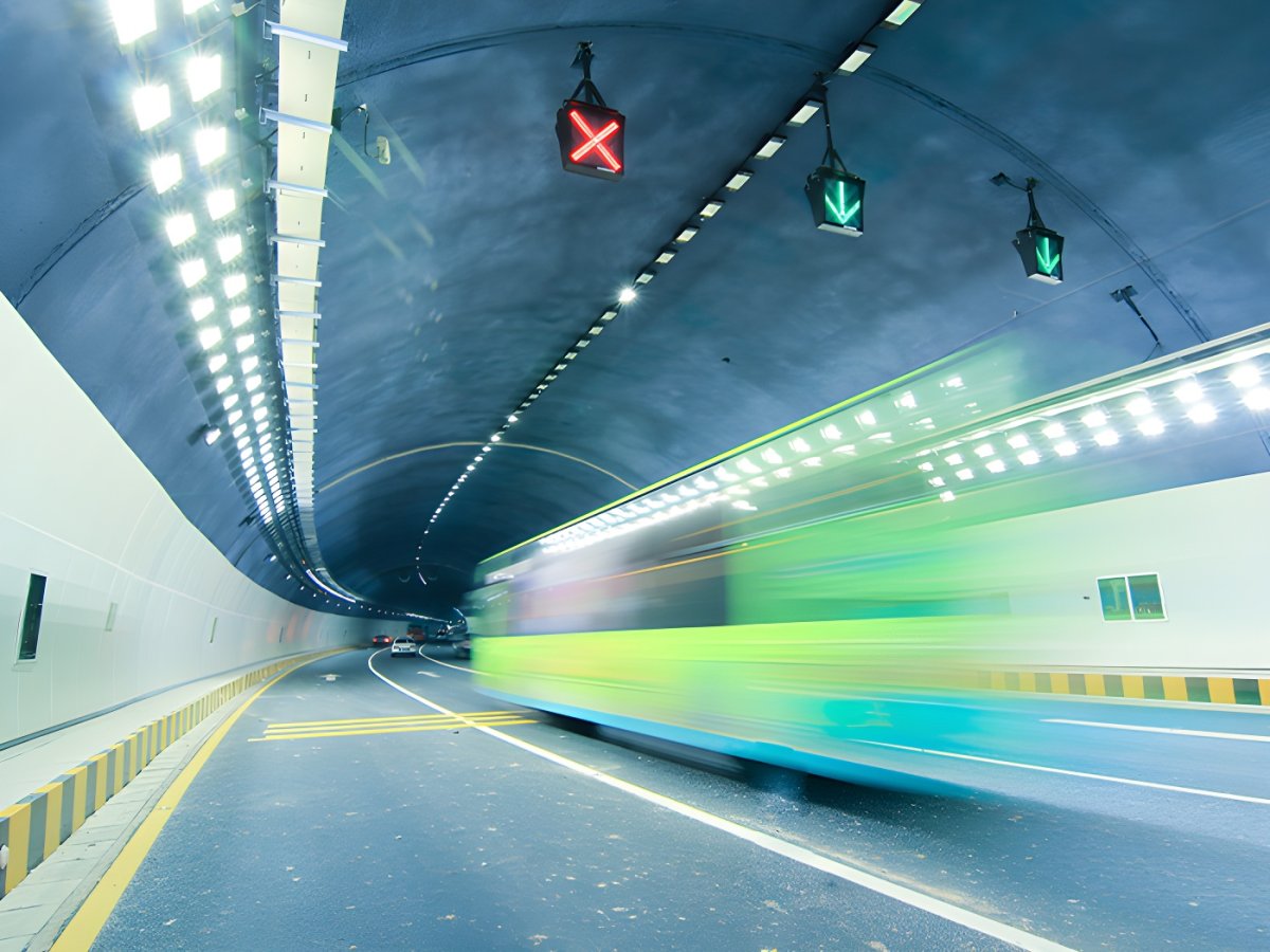 Motion blur of a green bus driving through a brightly lit highway tunnel with overhead digital lane control signals.