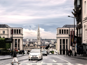 Cars and pedestrians on a Brussels street with the Town Hall spire visible in the distance under a cloudy sky.