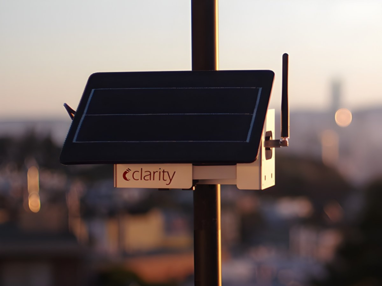 Clarity Movement air quality sensor with a solar panel and antenna mounted on a pole overlooking a city at sunset.