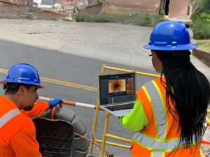 Two utility workers in hard hats and safety vests use a laptop to monitor a SewerAI pipe inspection on a city street.