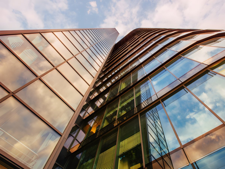 Low-angle view of a modern glass skyscraper reflecting a blue sky with white clouds.