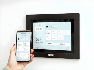 A hand holding a smartphone next to a wall-mounted Bisly smart building control panel displaying a digital interface.