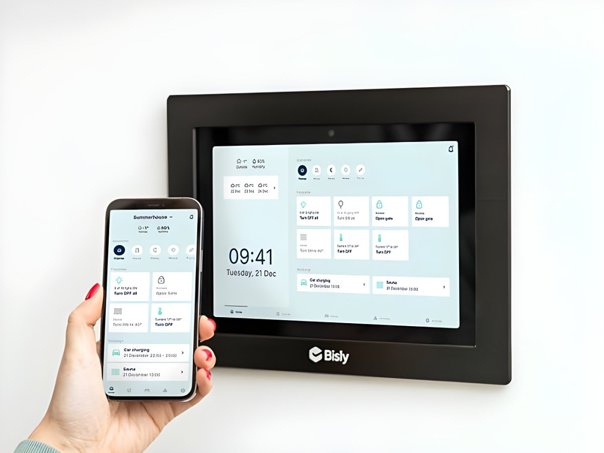 A hand holding a smartphone next to a wall-mounted Bisly smart building control panel displaying a digital interface.