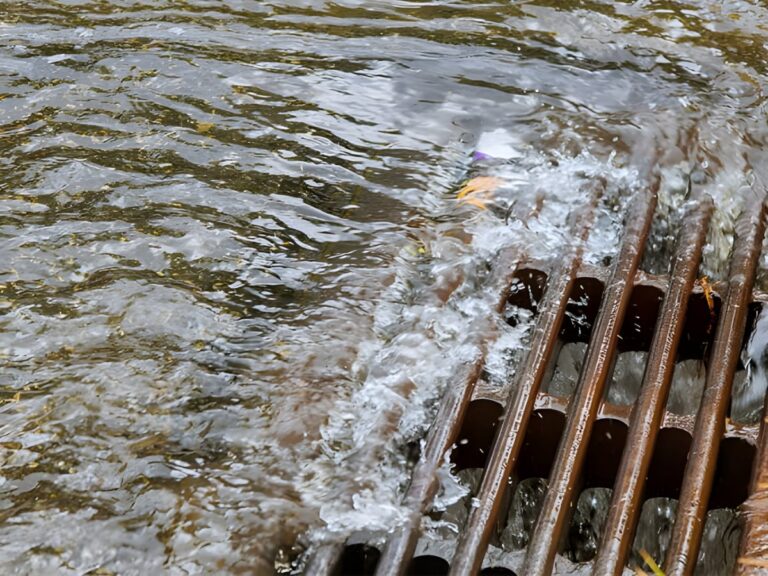 Water rushing through a rusted metal storm drain grate during a heavy rain event.