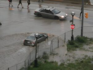 Cars driving through a flooded city intersection with pedestrians walking nearby during a heavy rainstorm.