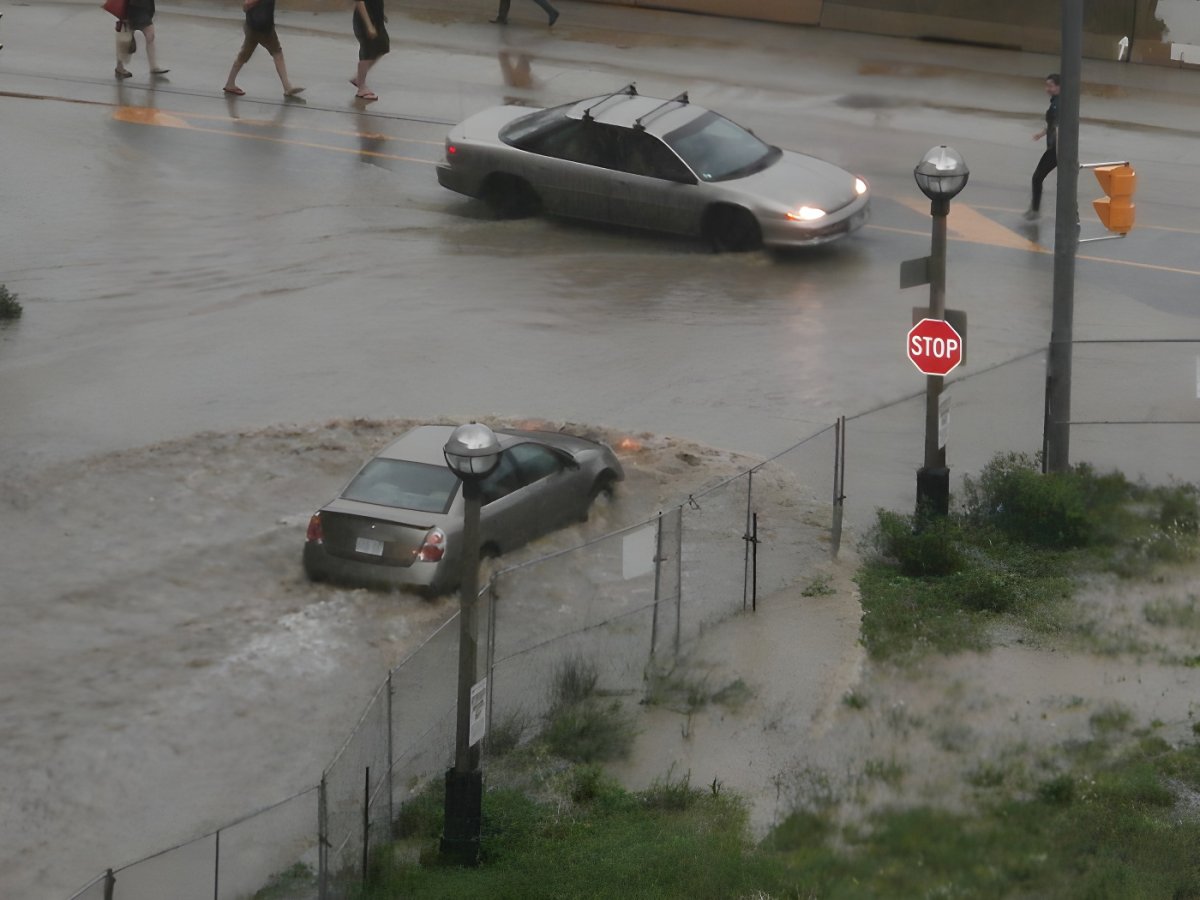 Cars driving through a flooded city intersection with pedestrians walking nearby during a heavy rainstorm.