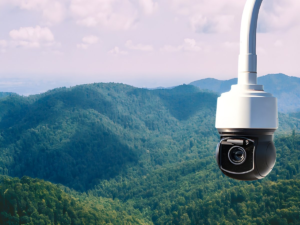 A white and black dome security camera mounted overlooking a vast, dense green forest and mountain range.