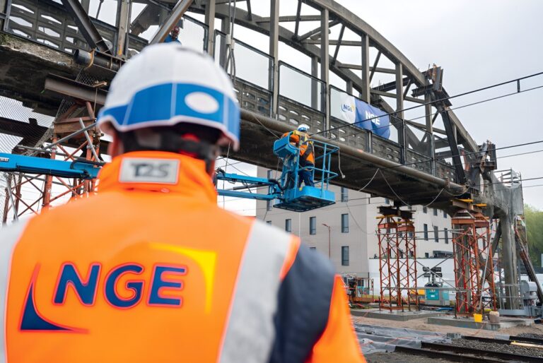 Worker in an NGE high-visibility vest watches a technician on a blue lift repairing a steel railway bridge.