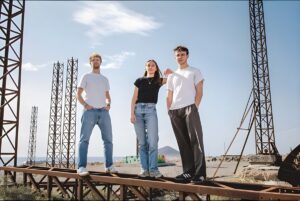 Three people standing on a rusted metal industrial frame outdoors with lattice towers and a clear sky in the background.