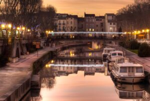 Illuminated street lamps and holiday lights reflect on the Canal de la Robine in Narbonne at dusk with moored canal boats.