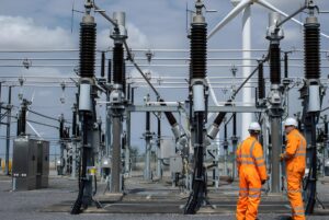 Two utility workers in orange high-visibility suits stand before high-voltage substation insulators and a wind turbine.