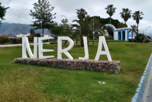 Large white Nerja sign on a stone base in a grassy park with palm trees and mountains in the background.