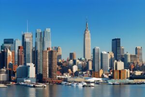 Midtown Manhattan skyline featuring the Empire State Building viewed from the Hudson River under a clear blue sky.