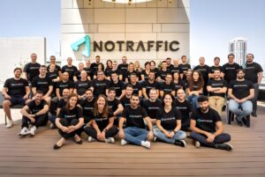 A large group of employees wearing branded t-shirts poses on a rooftop deck in front of a NoTraffic wall sign.