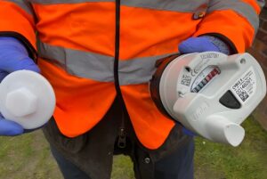 A technician in a high-visibility vest and blue gloves holds a Sensus smart water meter and a white plastic cap.