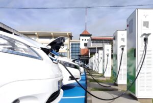 Row of white electric cars plugged into a line of tall charging stations in a paved parking lot.