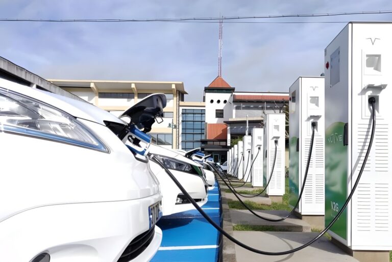 Row of white electric cars plugged into a line of tall charging stations in a paved parking lot.