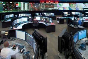 Operators at curved workstations monitor a large video wall displaying grid data and maps inside the CAISO control room.