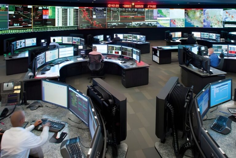 Operators at curved workstations monitor a large video wall displaying grid data and maps inside the CAISO control room.