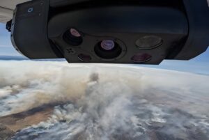 An Overwatch Imaging sensor mounted on an aircraft flying over a wildfire with thick white smoke.