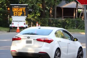 White Kia sedan driving past a portable digital road sign displaying the message UNSAFE STOP ! in a residential area.