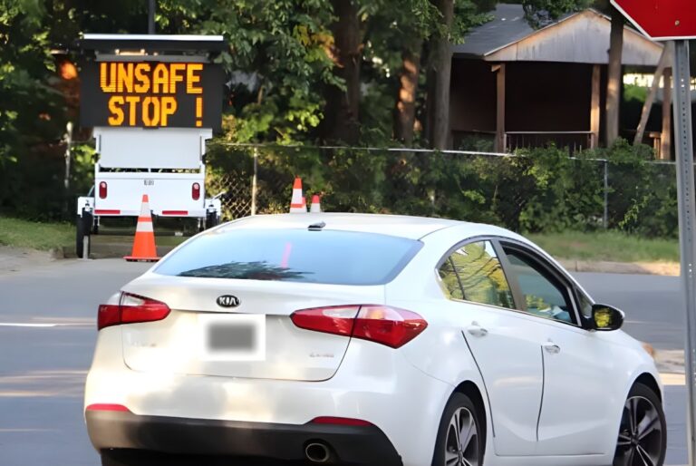 White Kia sedan driving past a portable digital road sign displaying the message UNSAFE STOP ! in a residential area.