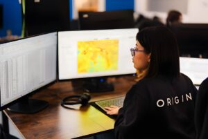 A person in an Origin Tech jacket views a digital heat map and data spreadsheets on dual monitors at a wooden desk.