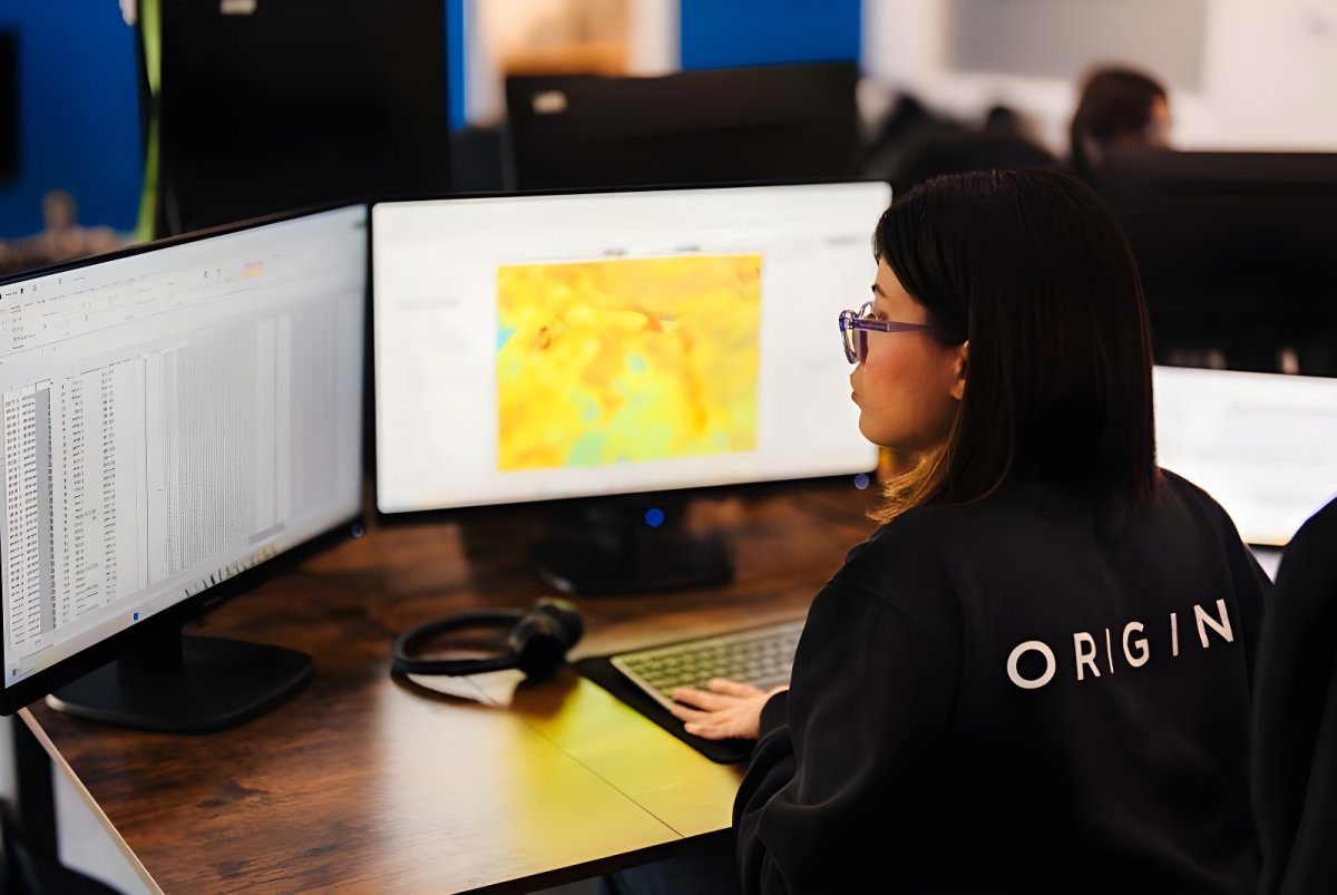 A person in an Origin Tech jacket views a digital heat map and data spreadsheets on dual monitors at a wooden desk.