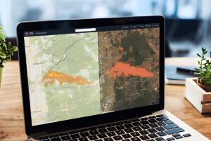 Laptop on a wooden desk displaying OroraTech wildfire monitoring software with split-screen satellite and topographic maps.