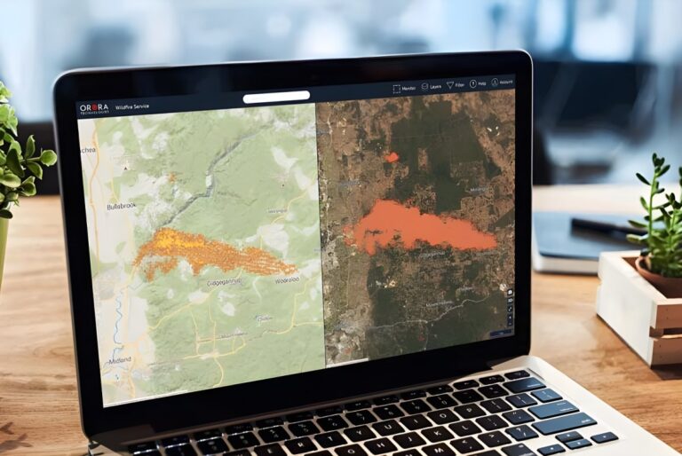 Laptop on a wooden desk displaying OroraTech wildfire monitoring software with split-screen satellite and topographic maps.