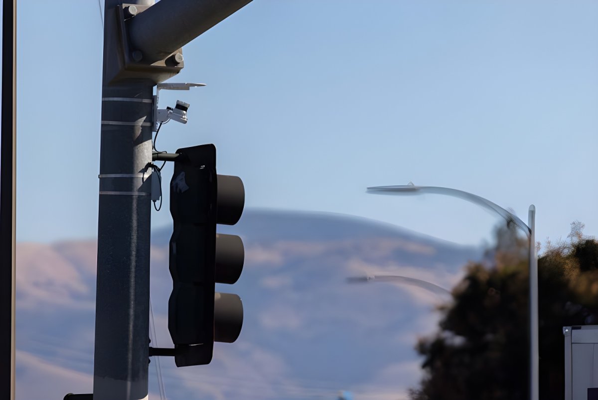 An Ouster lidar sensor mounted above a black traffic signal on a metal pole with hills in the background.
