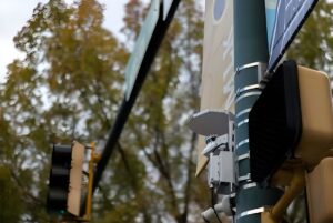 An Ouster lidar sensor mounted on a green traffic signal pole at an intersection with autumn trees in the background.