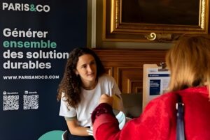 A woman at a Paris&Co information booth speaks with a visitor in front of a banner about sustainable solutions.