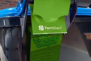 A green PermiServ tag hanging from the handle of a blue and black recycling bin.