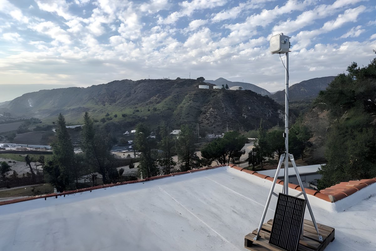 A QuantAQ air quality sensor mounted on a tripod with a solar panel on a rooftop overlooking hills in Pasadena.
