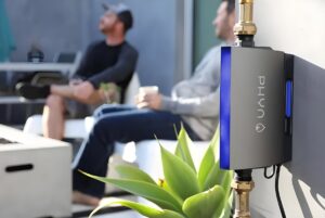 A Phyn smart water monitor installed on an exterior wall with two people sitting on a patio in the background.