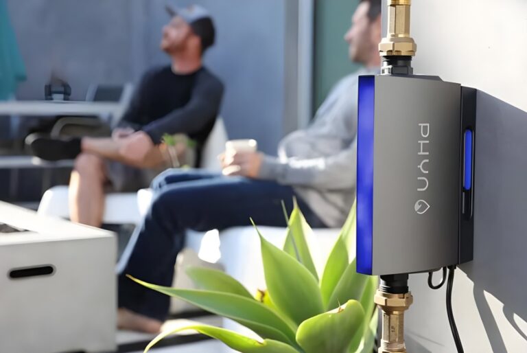 A Phyn smart water monitor installed on an exterior wall with two people sitting on a patio in the background.