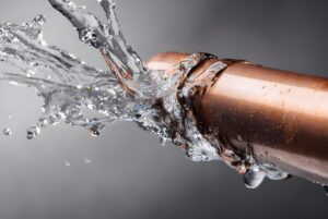 Water spraying and leaking from a fractured copper pipe against a gray background.