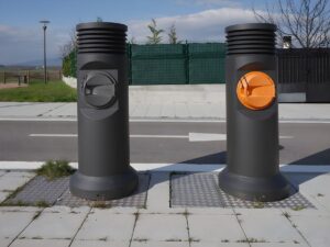 Two cylindrical pneumatic waste collection inlets on a sidewalk next to a paved road with a white directional arrow.